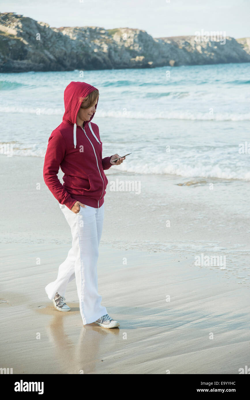 Young woman reading text message sur smartphone tout en flânant sur la plage, à Camaret-sur-mer, Bretagne, France Banque D'Images