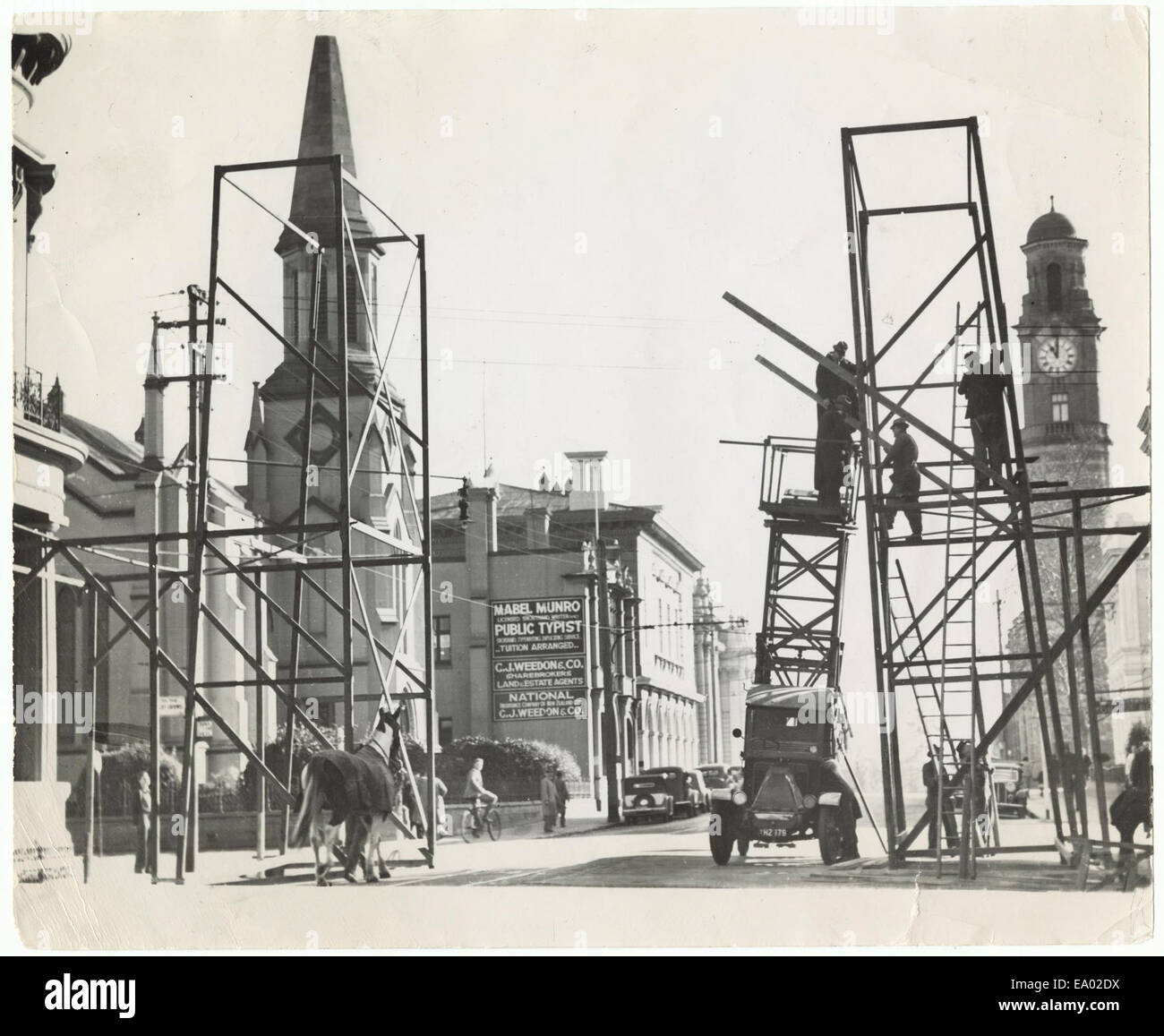 Cette photographie, prise par Burrows en 1946, montre une vue vers le nord le long de la rue St John à Launceston depuis le coin de la rue Paterson. Il capture les hommes qui construisent une arche V-Day, une célébration marquant la fin de la seconde Guerre mondiale en Europe, reflétant l'atmosphère d'après-guerre et les célébrations de la victoire dans la ville. Banque D'Images