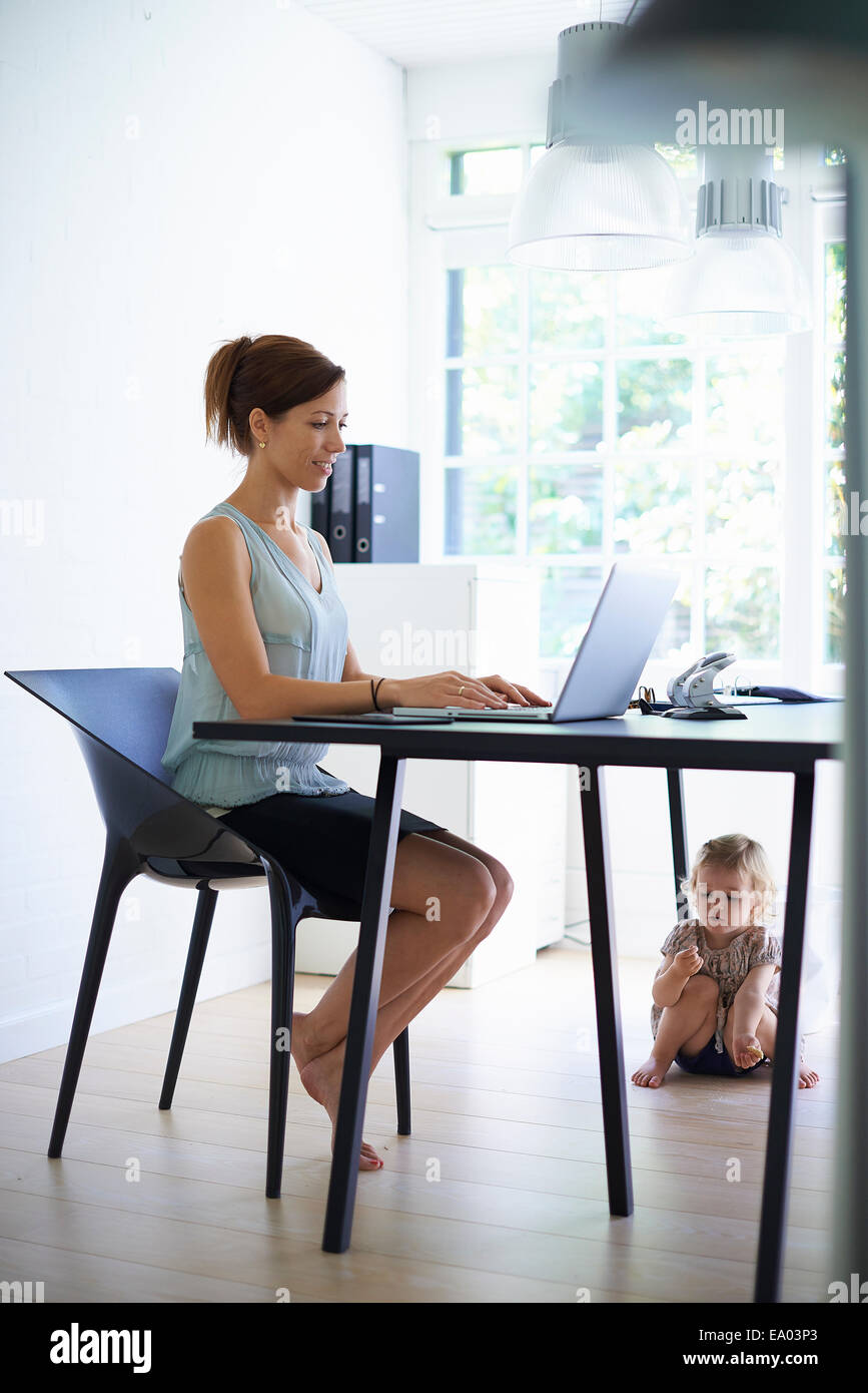 Mid adult mère saisie sur ordinateur portable avec toddler fille sous la table Banque D'Images