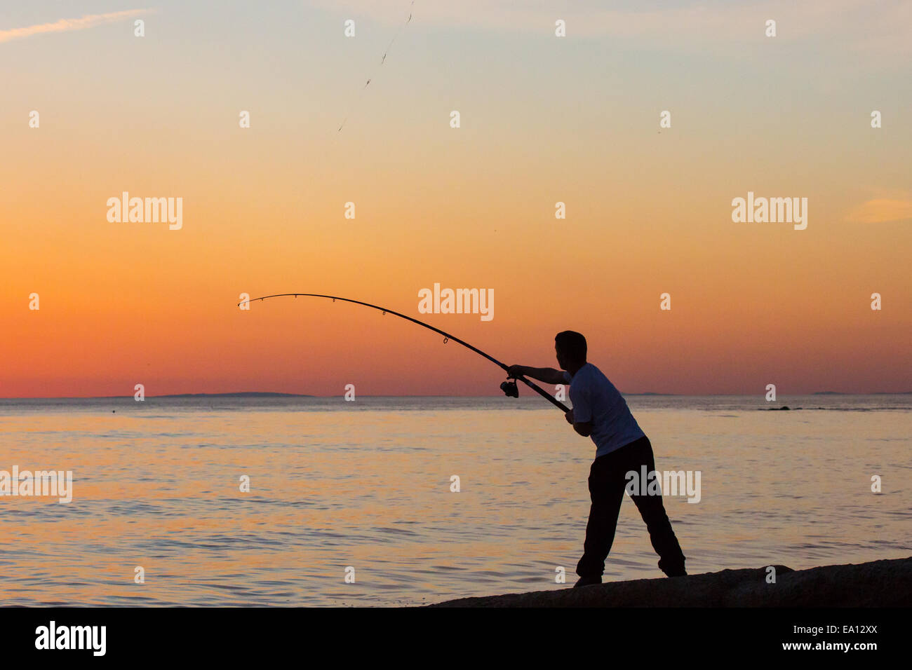 Un pêcheur solitaire sur le sihouetted terre contre un coucher de soleil orange Banque D'Images