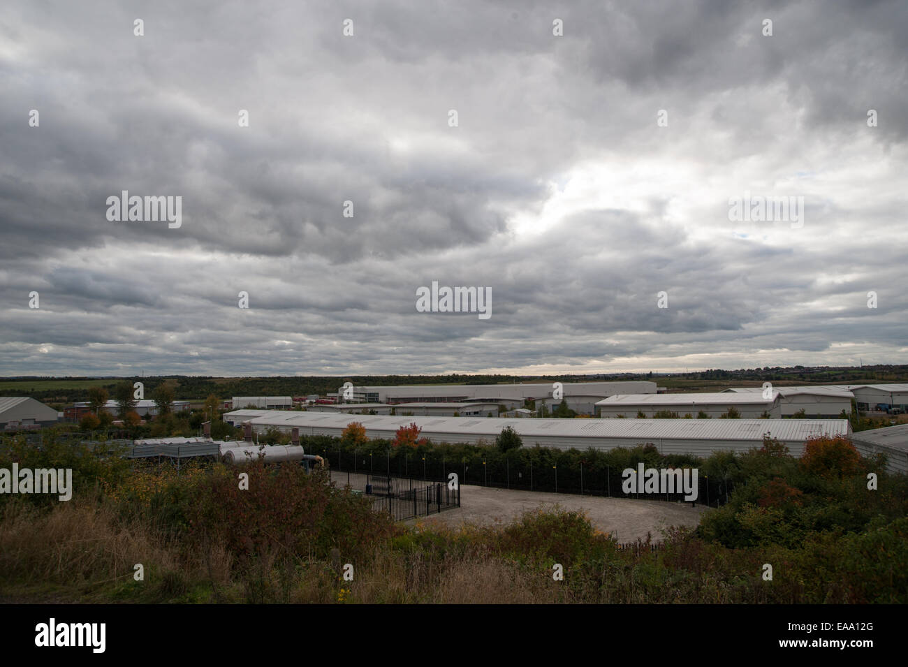 Vue large des entrepôts industriels dans un paysage rural, avec des logements lointains et un ciel nuageux sur une zone à usage mixte. Banque D'Images