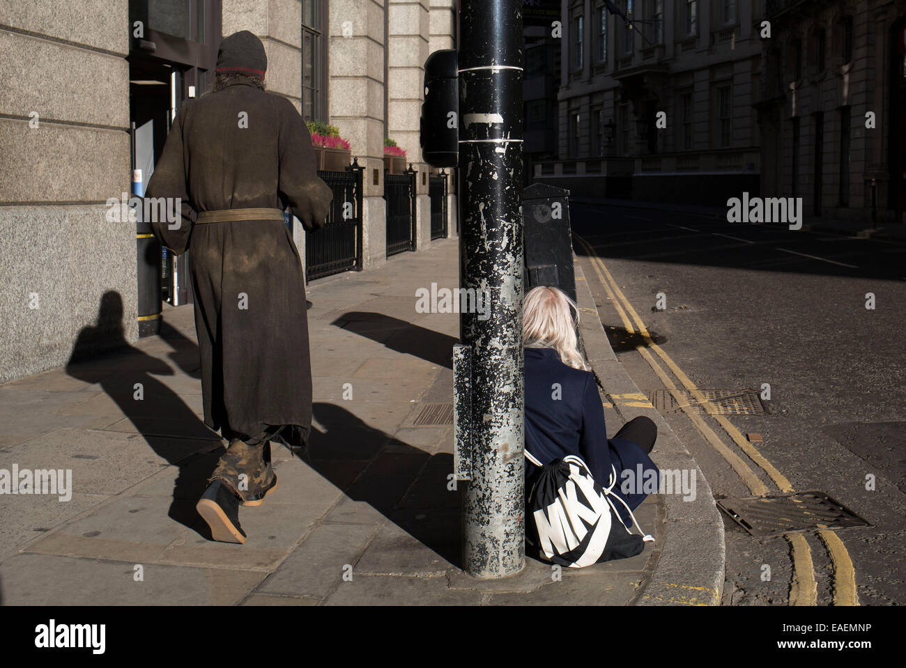 Un sans-abri passe devant une fille qui est de compter le nombre de personnes qui passent dans la rue. Elle est assise sur le sol de sa collecte Banque D'Images