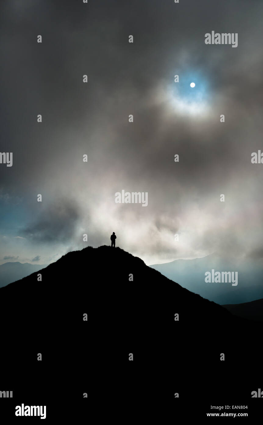 Un homme (uniquement d'ossature visible) debout sur un sommet de montagne / peak. Au-dessus de lui le soleil peut être vu briser les nuages. Banque D'Images