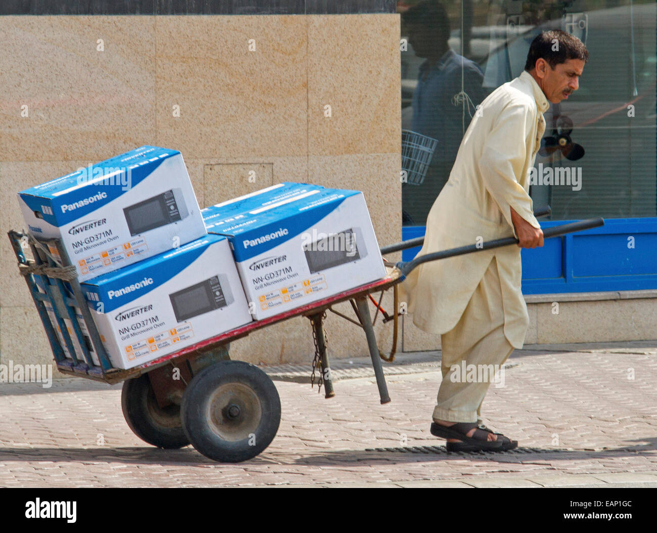 L'homme asiatique, travailleur en costume traditionnel tirant barrow / chariot chargé de boîtes avec des biens de consommation électroniques le long de rue de ville à Dubaï ÉMIRATS ARABES UNIS Banque D'Images