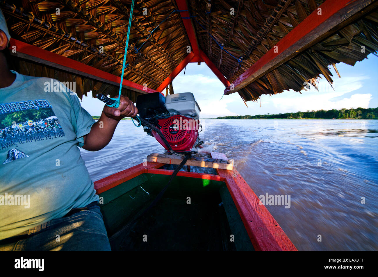 Un chauffeur de taxi de l'eau steers son moteur hors-bord le long d'une rivière au coucher du soleil passé un rainforest berge. Banque D'Images