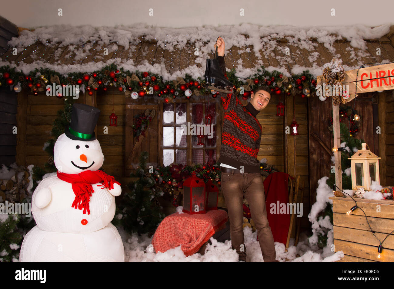 Jeune homme blanc Holding Paire de patins à glace jusqu'à la maison en bois décorés avec Bonhomme Hiver et Noël Décors Booth. Banque D'Images