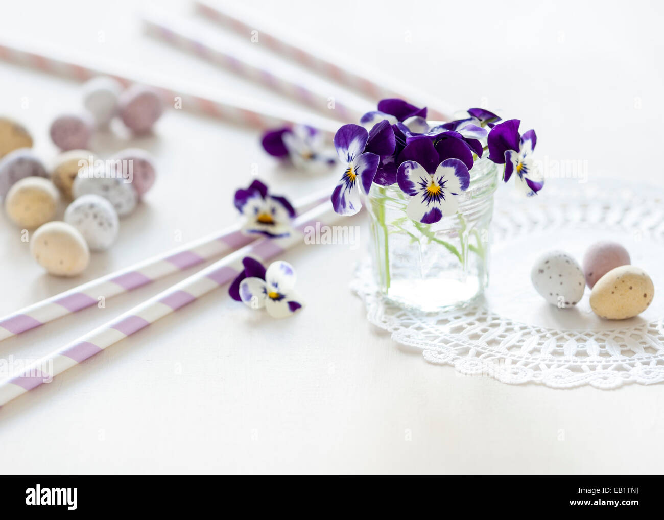 Petit bouquet de pensées dans un bocal en verre sur un napperon sur le tableau blanc, avec des mini-oeufs et pailles pastel tous baigné de soleil Banque D'Images