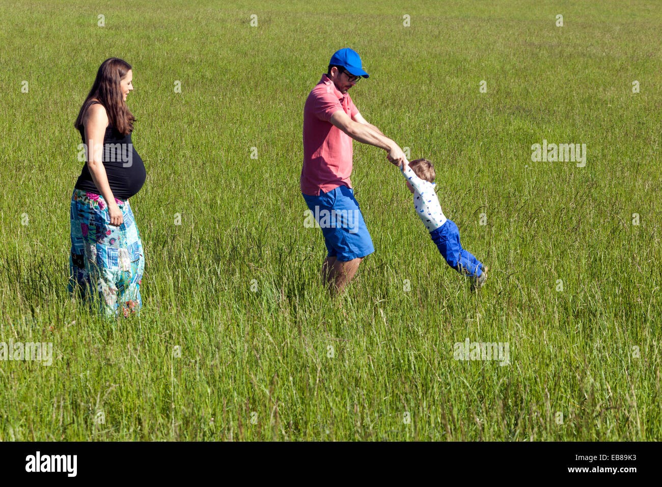 Une famille heureuse, une femme enceinte à pied avec son mari et son petit garçon passer du temps ensemble et de jouer à des jeux dans une prairie d'été Banque D'Images