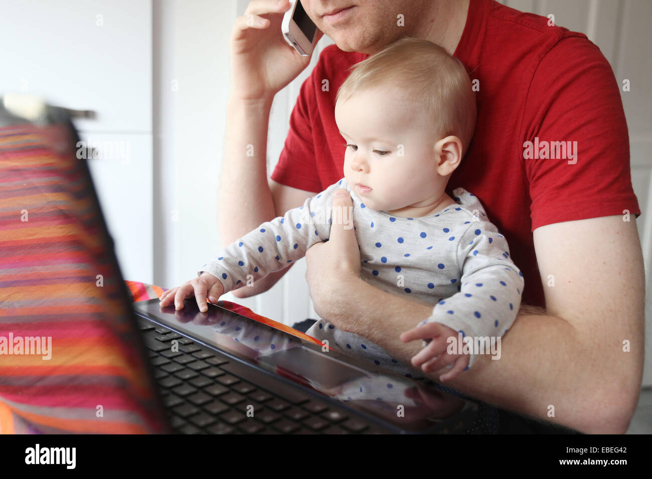 Un père travaille sur son ordinateur portable à partir de la maison tout en tenant son bébé de huit mois. Banque D'Images