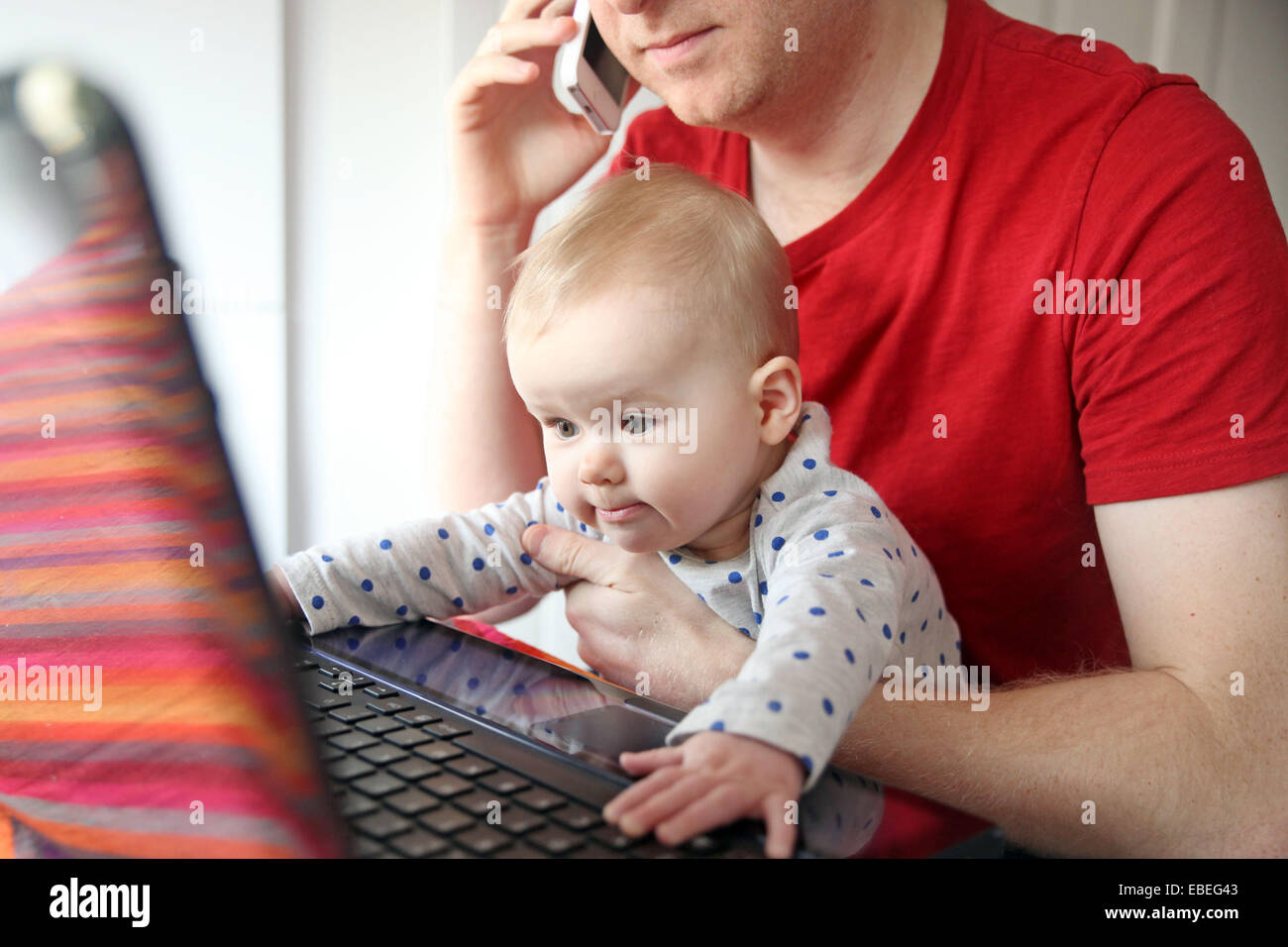 Un père travaille sur son ordinateur portable à partir de la maison tout en tenant son bébé de huit mois. Banque D'Images