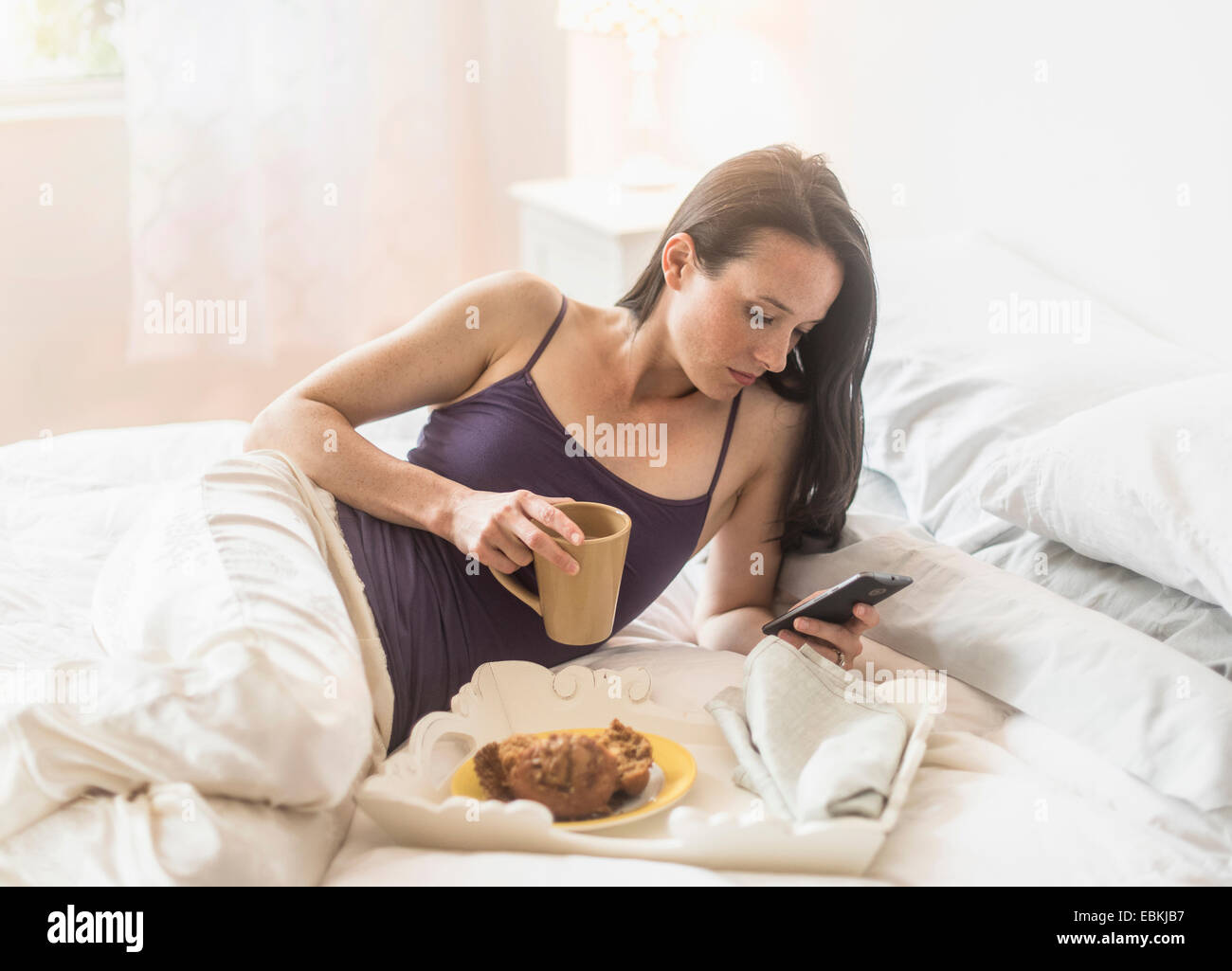 Woman having breakfast in bed, using mobile phone Banque D'Images