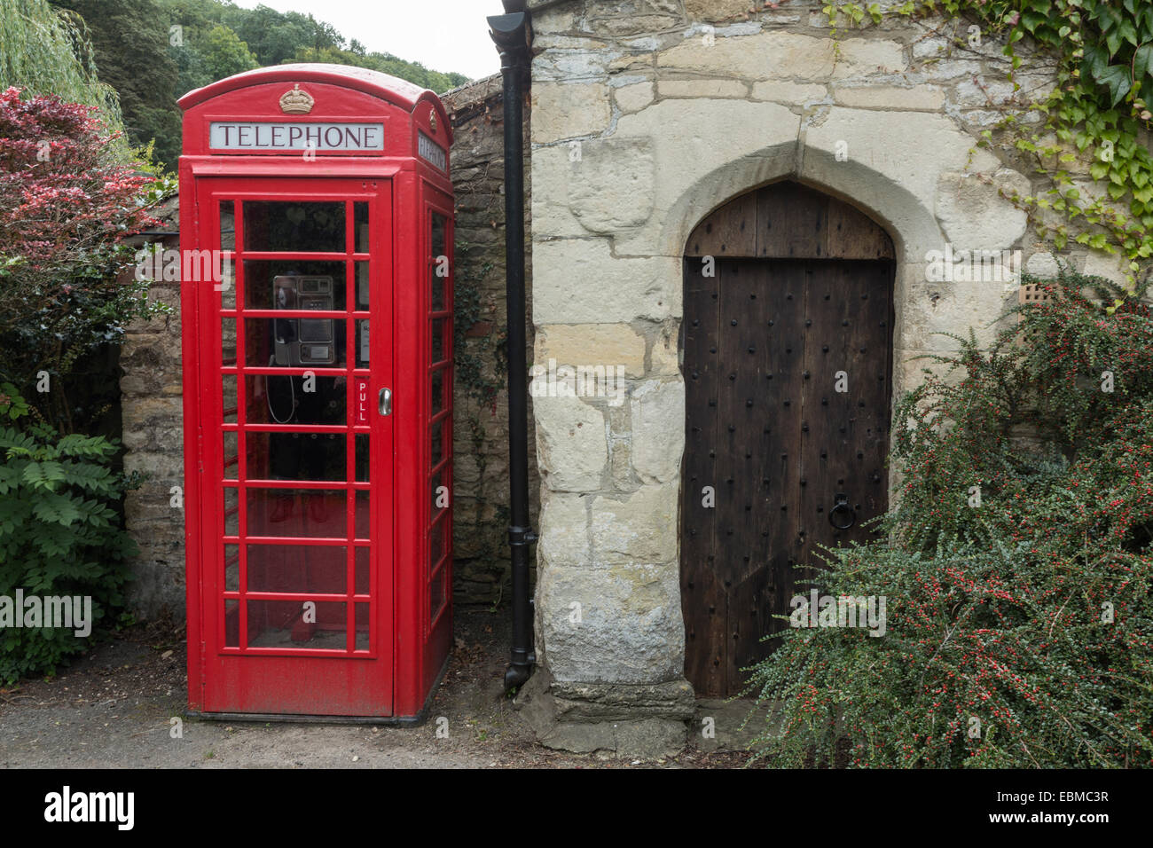 Téléphone rouge case à côté d'une vieille porte en bois cloutée dans un bâtiment en pierre de Cotswold, Castle Combe, Wiltshire, Angleterre Banque D'Images