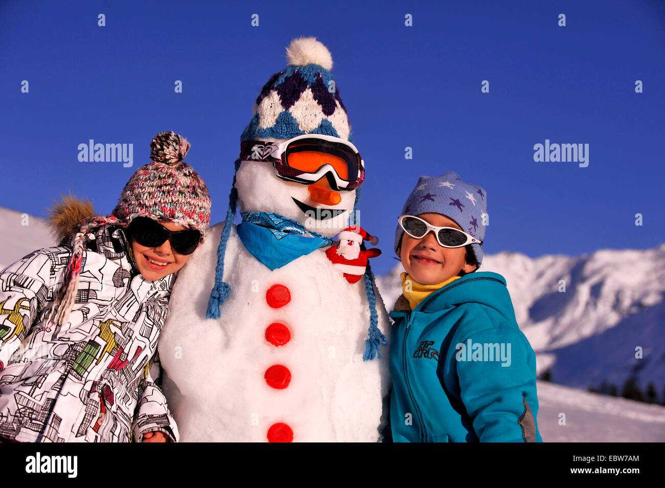 Deux jeunes garçons à côté d'un bonhomme avec bobble hat et lunettes de ski en face de montagnes couvertes de neige et ciel bleu, France Banque D'Images