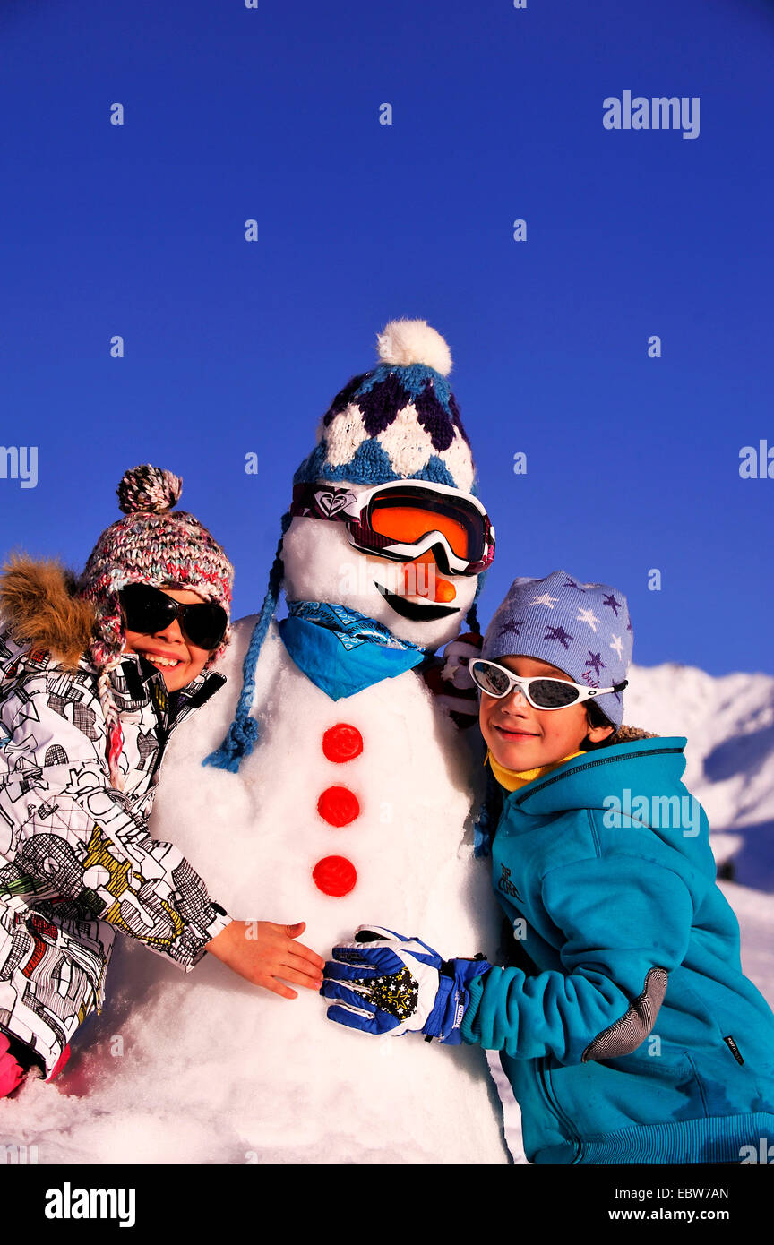 Deux jeunes garçons à côté d'un bonhomme avec bobble hat et lunettes de ski en face de montagnes couvertes de neige et ciel bleu, France Banque D'Images