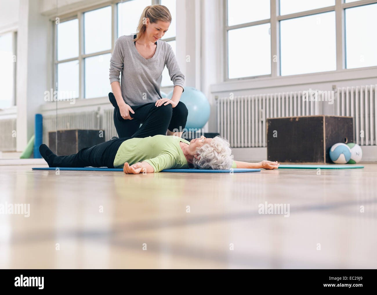 Femme thérapeute physique d'aider les femme avec exercice des jambes dans la thérapie de sport. Elder woman lying on floor étant assisté par Banque D'Images Femme thérapeute physique d'aider les femme avec exercice des jambes dans la thérapie de sport. Elder woman lying on floor étant assisté par Banque D'Images