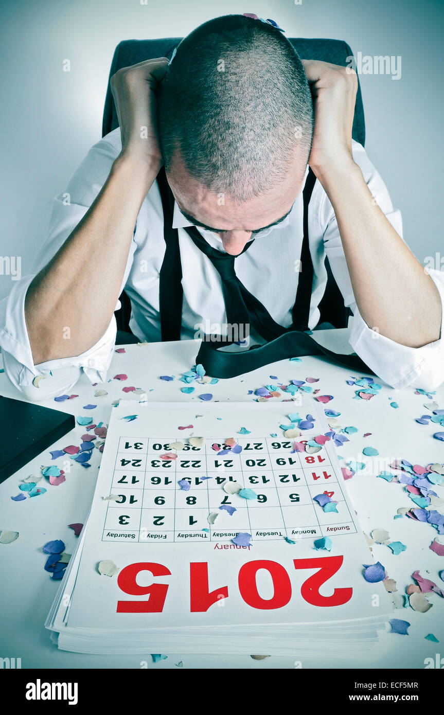 Un jeune homme dans le bureau avec sa tête dans ses mains après un nouveau ans parti, avec un calendrier de l'année 2015 et les confettis sur son bureau Banque D'Images