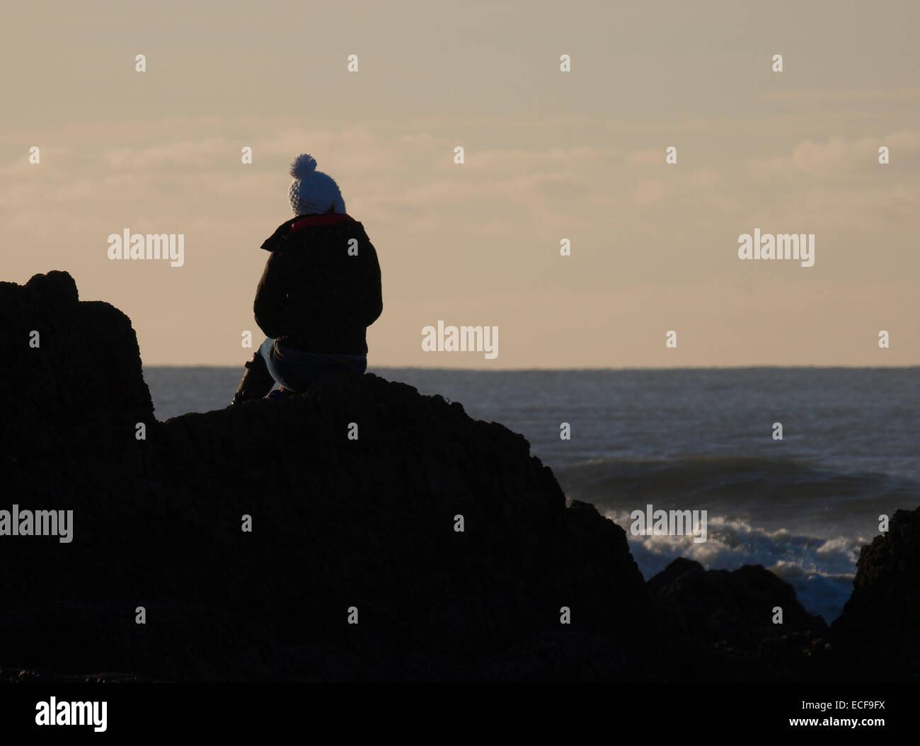 Teenage girl wearing a bobble hat assis sur des rochers surplombant la mer, Bude, Cornwall, UK Banque D'Images