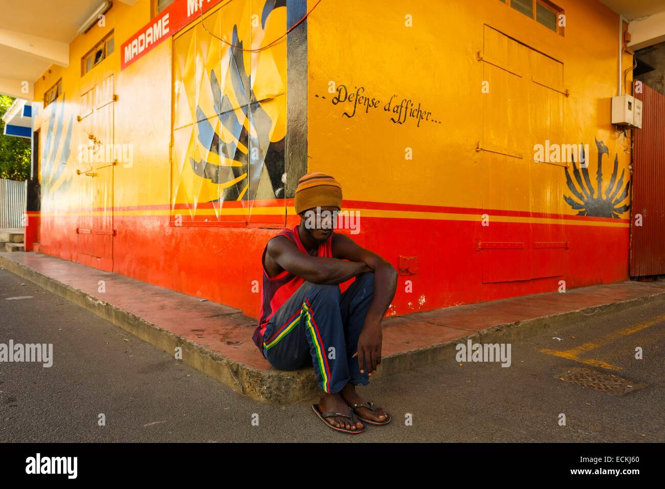 L'Île Maurice, Rodrigues, Port Mathurin, rasta créole homme assis sur le trottoir à l'angle d'une rue de la publicité avant une marque de spiritueux Banque D'Images