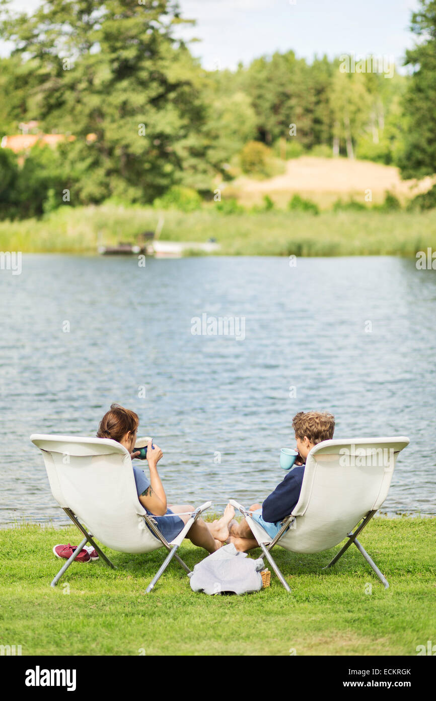 Vue arrière du couple having coffee sur les chaises longues au bord du lac de chaises Banque D'Images Vue arrière du couple having coffee sur les chaises longues au bord du lac de chaises Banque D'Images