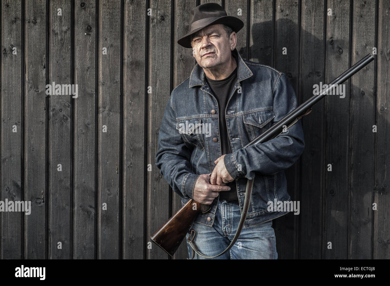 L'homme à chapeau et veste jeans avec un fusil devant un mur en regardant la caméra. Banque D'Images