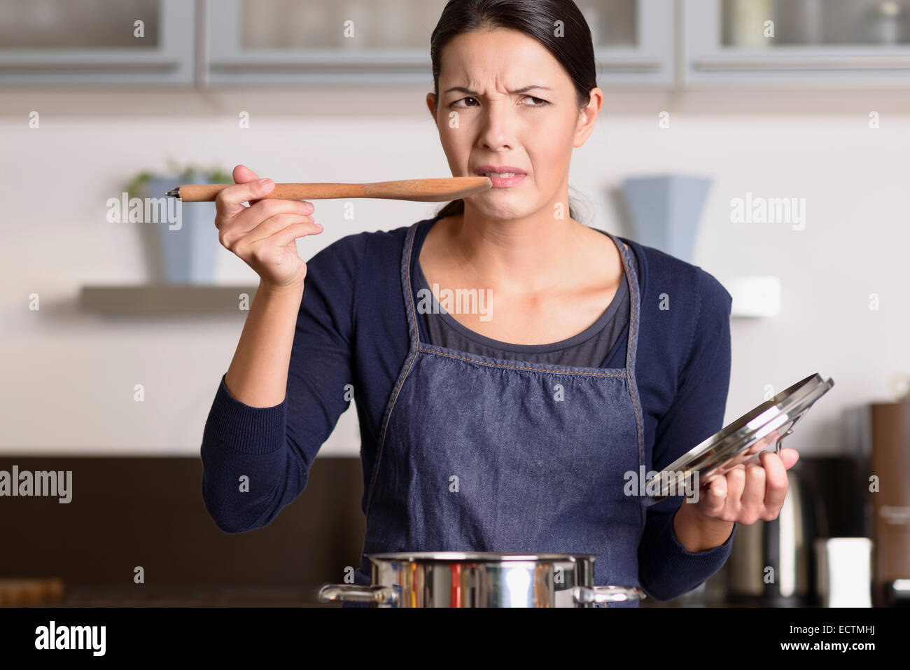 Jolie jeune femme cuisinière debout à la plaque de cuisson dans son tablier dégustation sa nourriture dans la casserole avec une grimace alors qu'elle est d'avis qu'il est di Banque D'Images