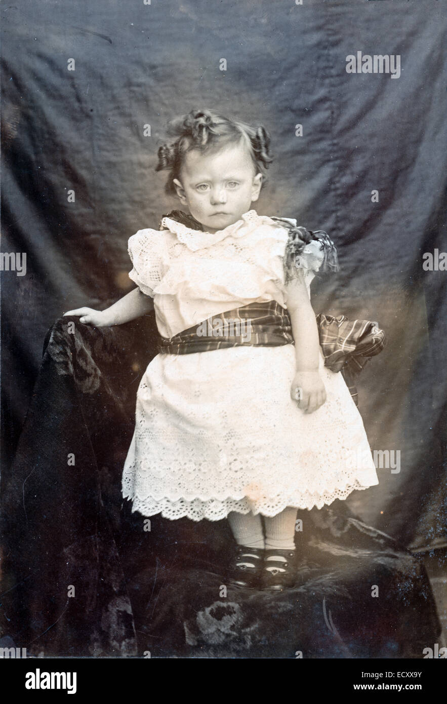 Photo en noir et blanc vers 1900. Studio portrait d'un garçon de deux ans à l'aspect malheureux debout dans sa robe de baptême avec un arrière-plan en tissu. Banque D'Images