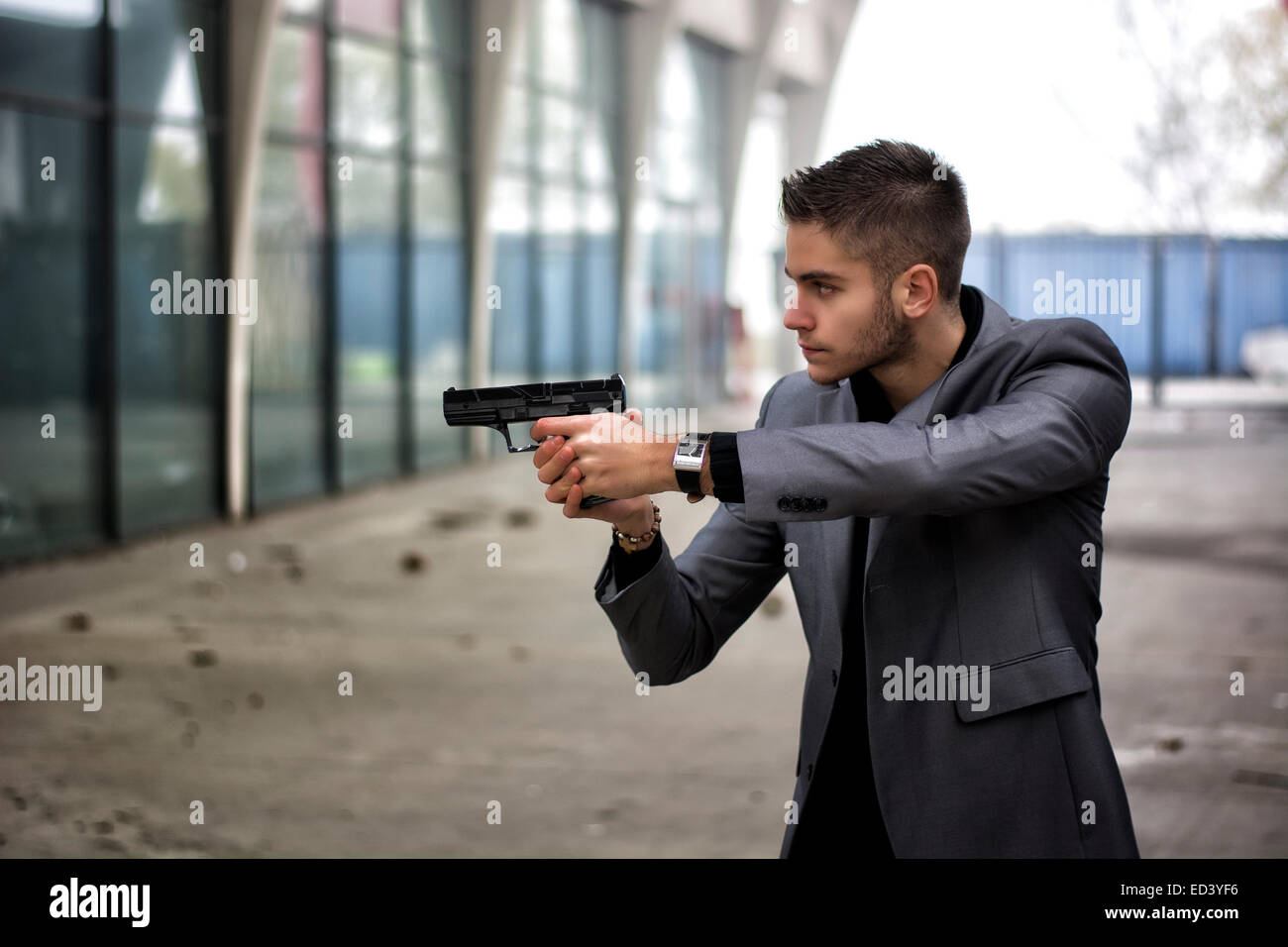 Bien habillé beau jeune détective ou policier ou gangster debout dans un environnement urbain visant une arme à feu sur la gauche Banque D'Images