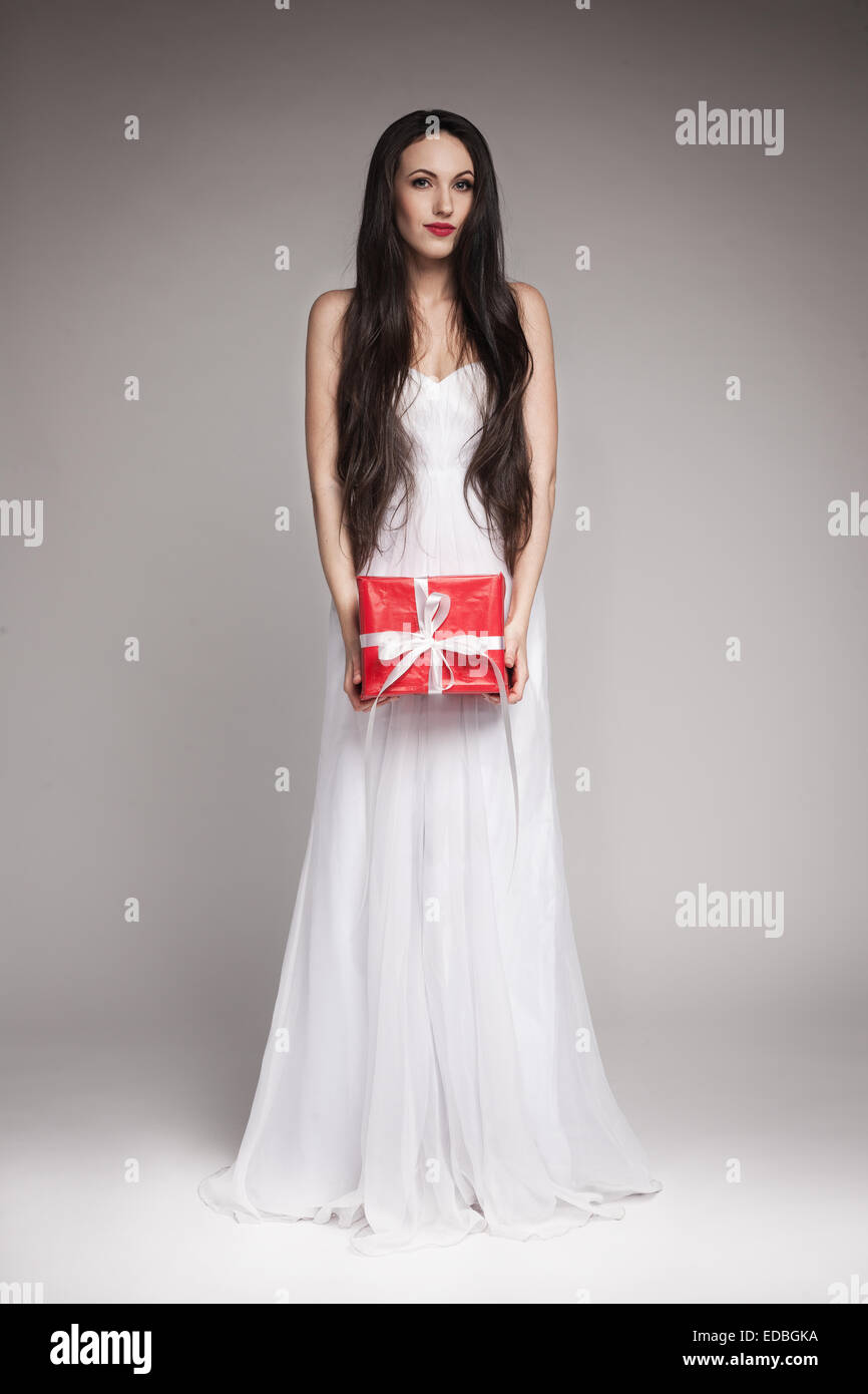 Belle jeune femme aux longs cheveux noirs portant des maxi robe blanche, holding gift enveloppée de papier rouge avec ruban blanc. Banque D'Images