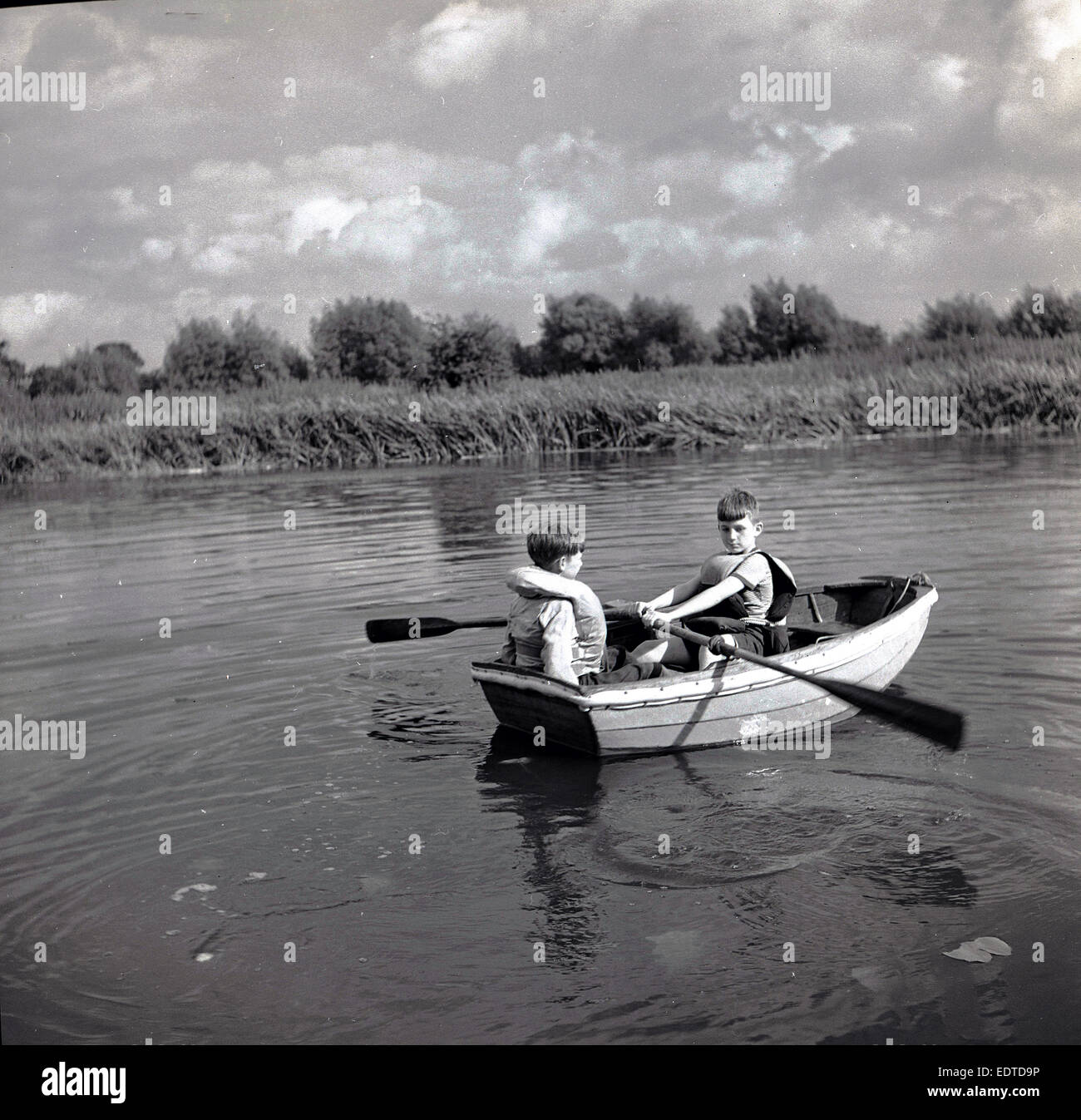 Années 1950, tableau historique, deux jeunes garçons portant des gilets de sauvetage d'un petit bateau à rames sur une rivière calme. Banque D'Images