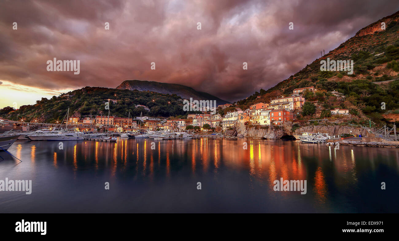 Coucher de soleil sur le port de Maratea, avec les derniers rayons de soleil stick out par la couleur rouge de nuages sur la ville, tandis que la première des lumières de rue se reflètent dans la mer. Tramonto al porto di Maratea, con montrer les raggi di sole amplificati da una nuvola sopra la città, mentre le premier Luci della strada nel mare riflettono le si. Banque D'Images