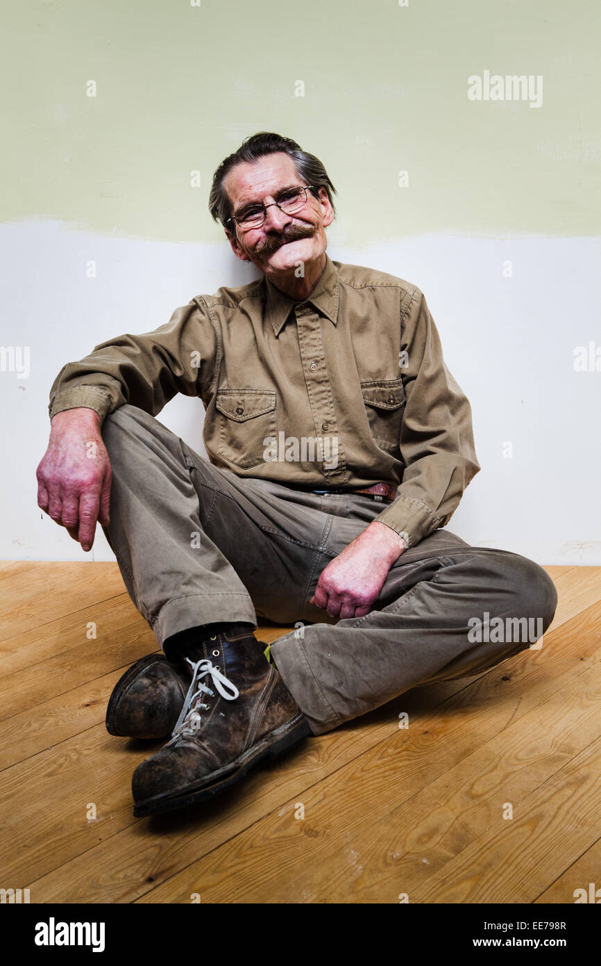 L'homme dans son 60s avec moustache portant chemise et pantalon kaki assis sur le plancher. Banque D'Images