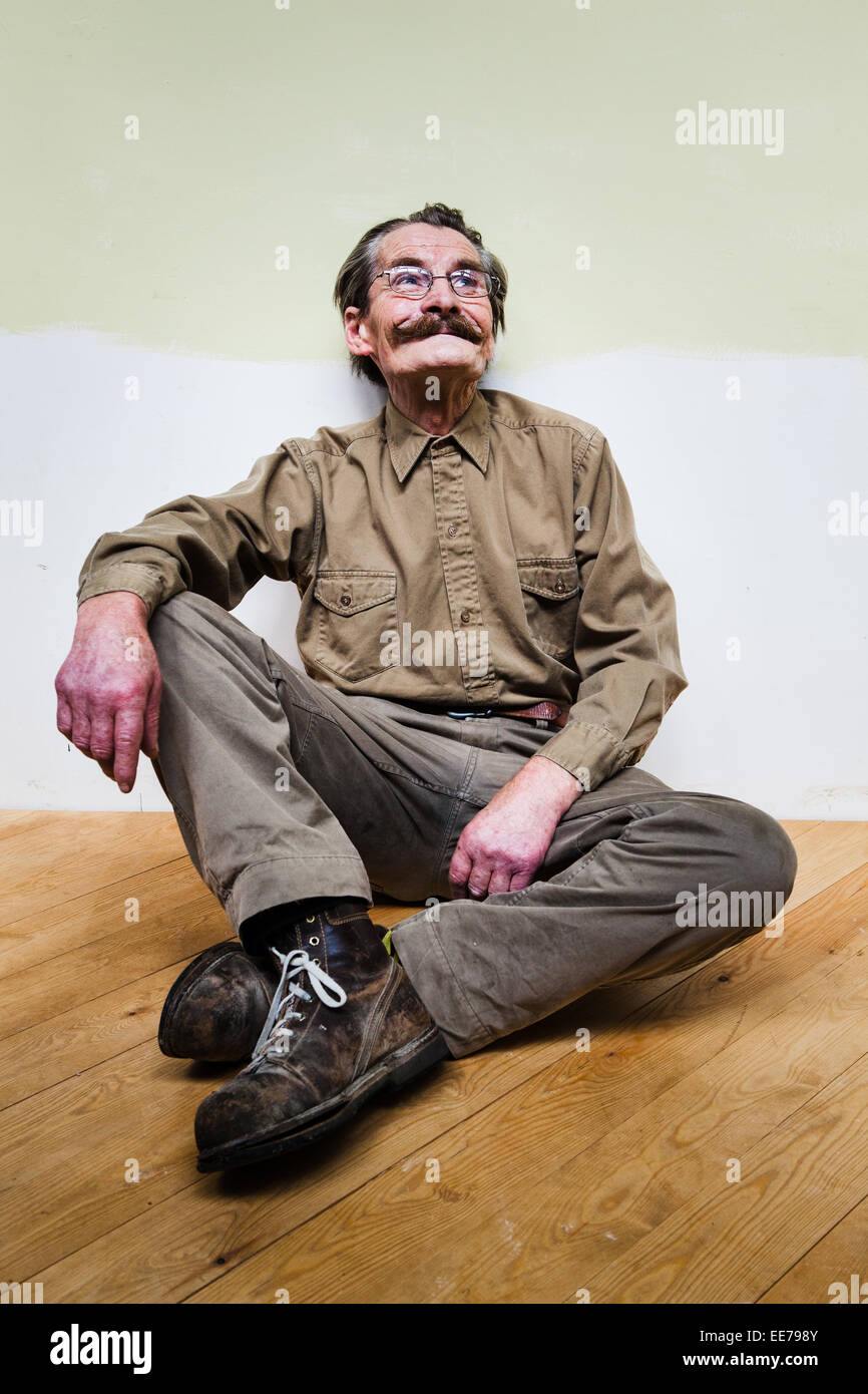 L'homme dans son 60s avec moustache portant chemise et pantalon kaki assis sur le plancher. Banque D'Images