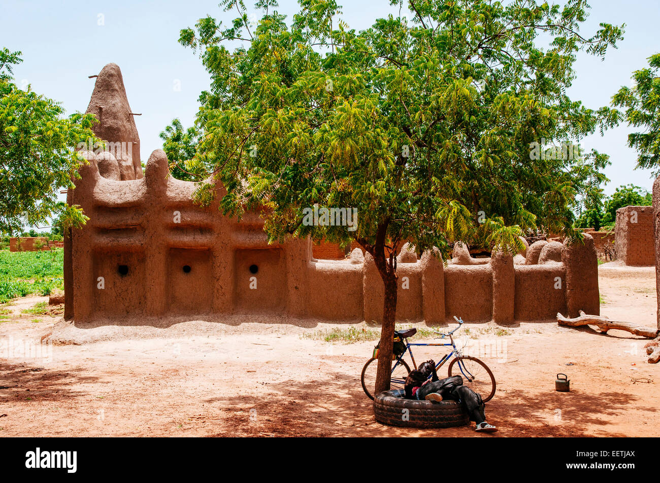 Jeune homme se reposant dans l'ombre d'un arbre juste en face d'une mosquée de style sahélien. Le Burkina Faso. Banque D'Images