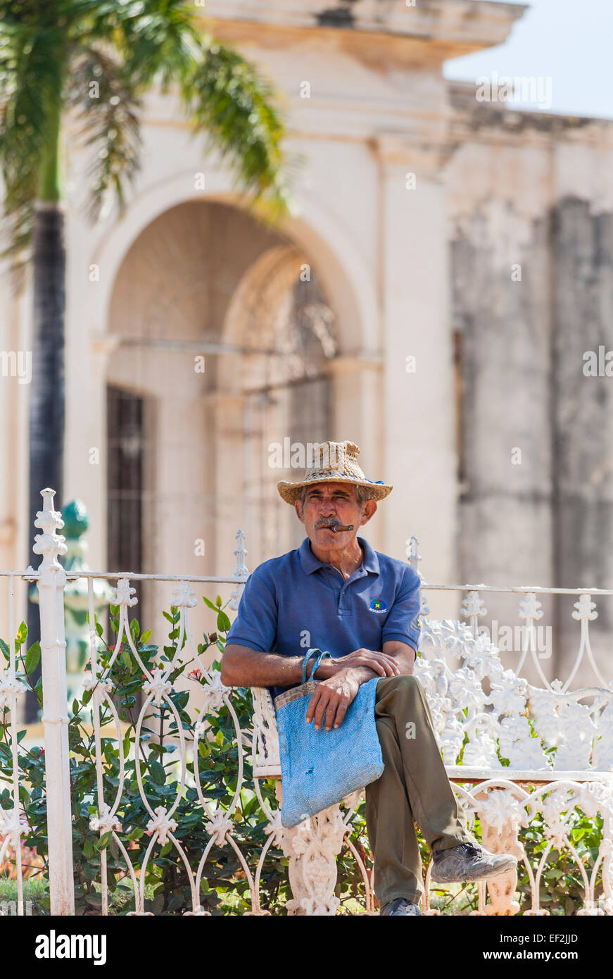 Style de vie cubain : homme d'âge moyen local avec une moustache portant un  chapeau de paille de détente assis sur un banc, fumant un cigare cubain, le  centre-ville de Trinidad, Cuba