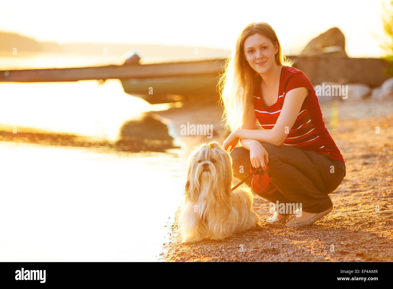 Jeune femme avec shih-tzu chien assis sur le bord du lac. La lumière de soleil rouge. Banque D'Images