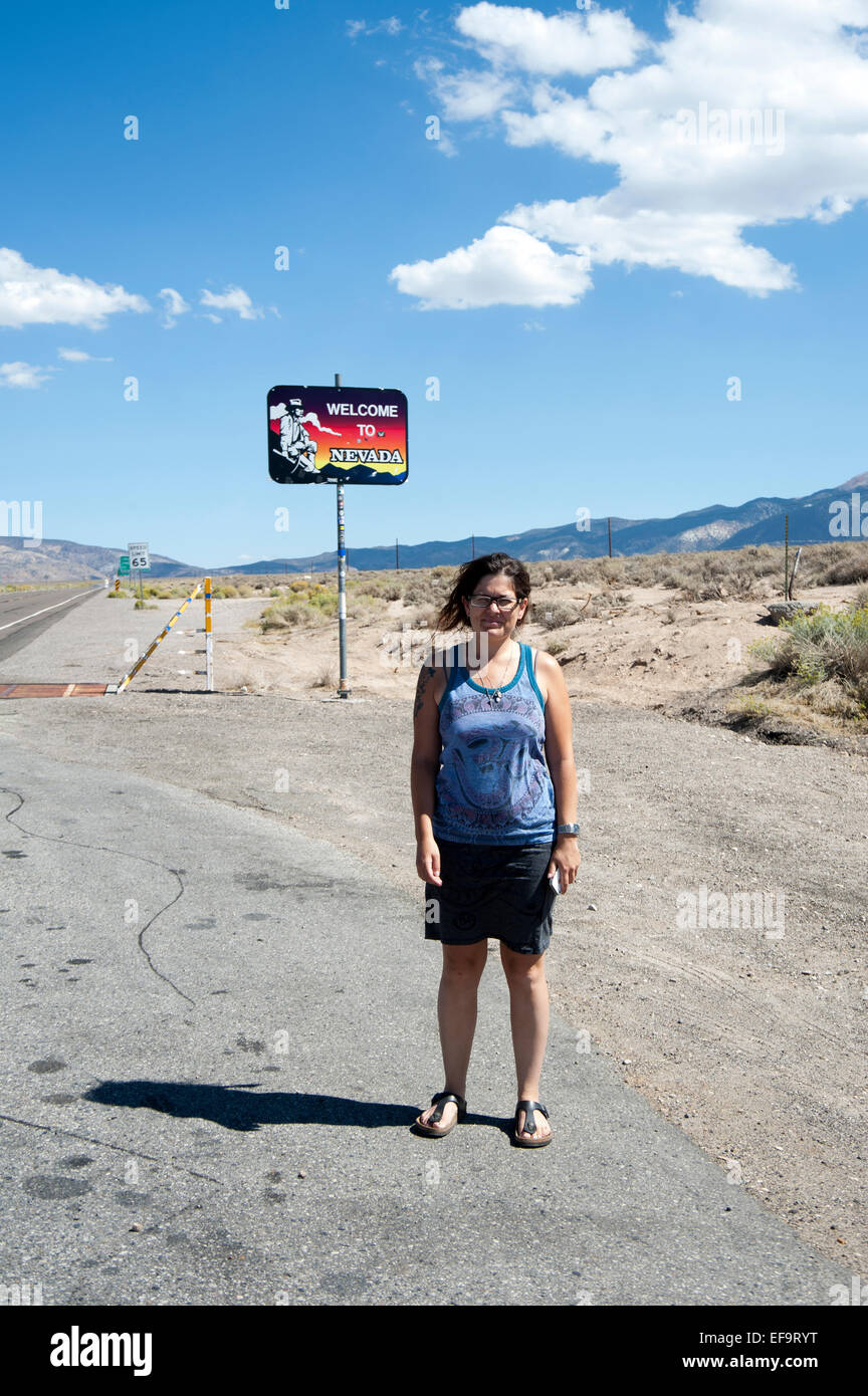 Femme debout dans un paysage aride au bord de l'est de la Californie avec un signe indiquant bienvenue au Nevada, USA Banque D'Images