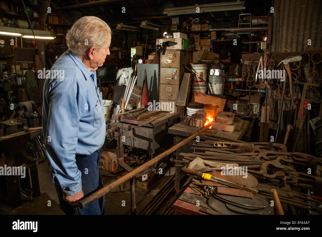 Un vieux forgeron à la vieille ville district de Tustin, CA CHAUFFE, une barre de fer rouge la forge de son atelier. Outils de notes sur l'établi. Banque D'Images