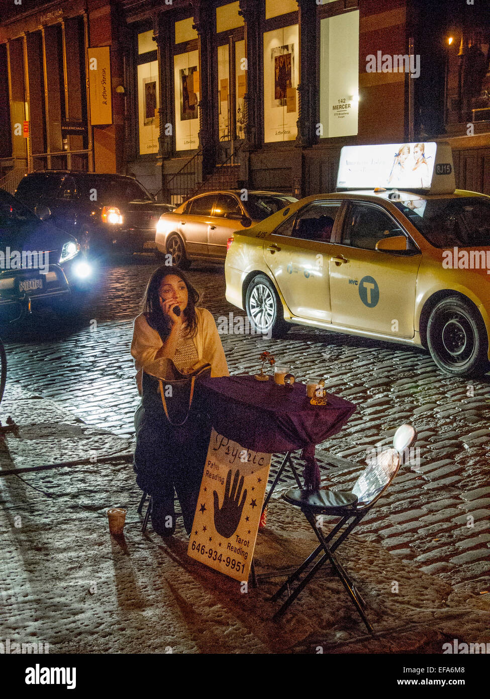 Un trottoir et psychiques tarot reader parle au téléphone cellulaire pendant l'attente pour les clients à une table pliante sur un trottoir de Manhattan de nuit à Soho, New York City. Note des pavés et taxi. Banque D'Images
