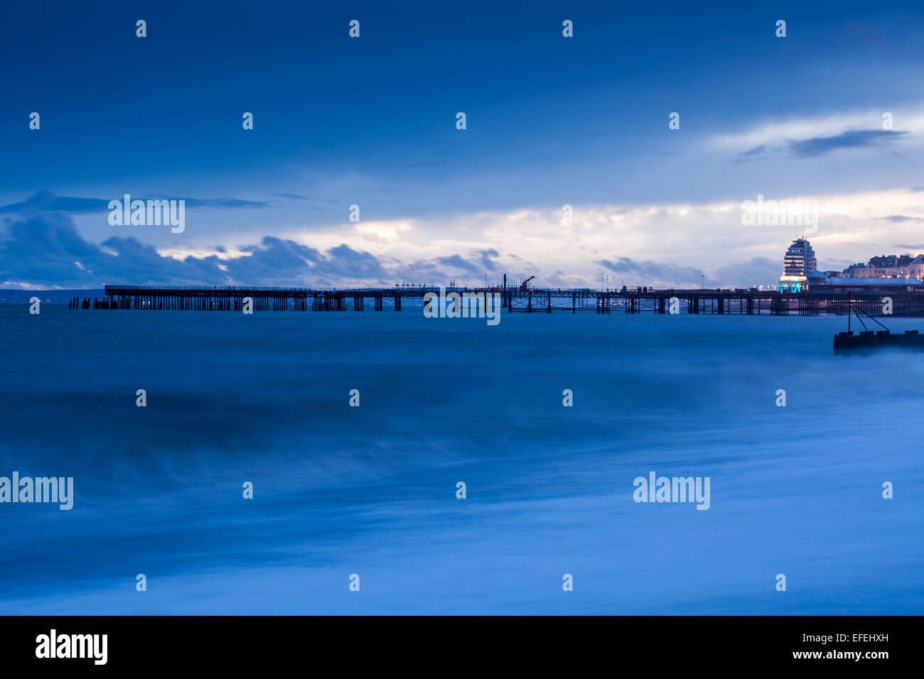 Front de mer de Hastings et de la jetée. Les vagues déferlent sur la plage au coucher du soleil. alors que les lumières de la ville scintillent dans l'arrière-plan. Banque D'Images