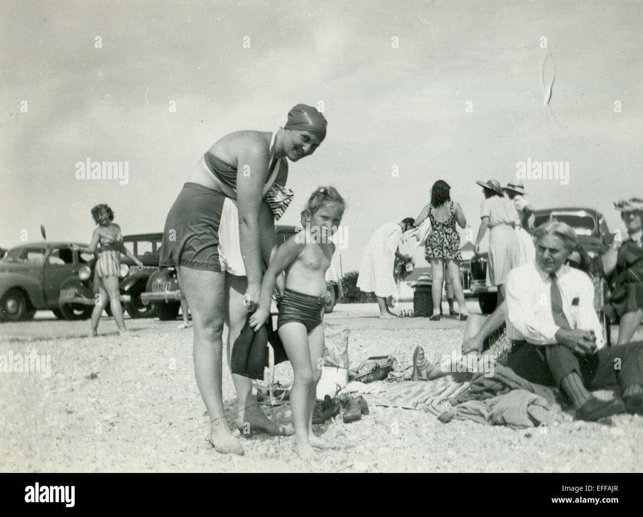 Le 12 décembre 2014 - Canada - circa 1930 : Reproduction d'une ancienne photo montre une femme en maillot de bain et d'un capuchon en caoutchouc mesure une petite fille sur la plage, regarder les personnes âgées businessman dans une égalité, pas de veste, qui se trouve à côté du sable. Dans l'arrière-plan une femme en maillot et une robe et un chapeau contre la toile de voitures. (Crédit Image : © Igor Golovniov/ZUMA/ZUMAPRESS.com) fil Banque D'Images