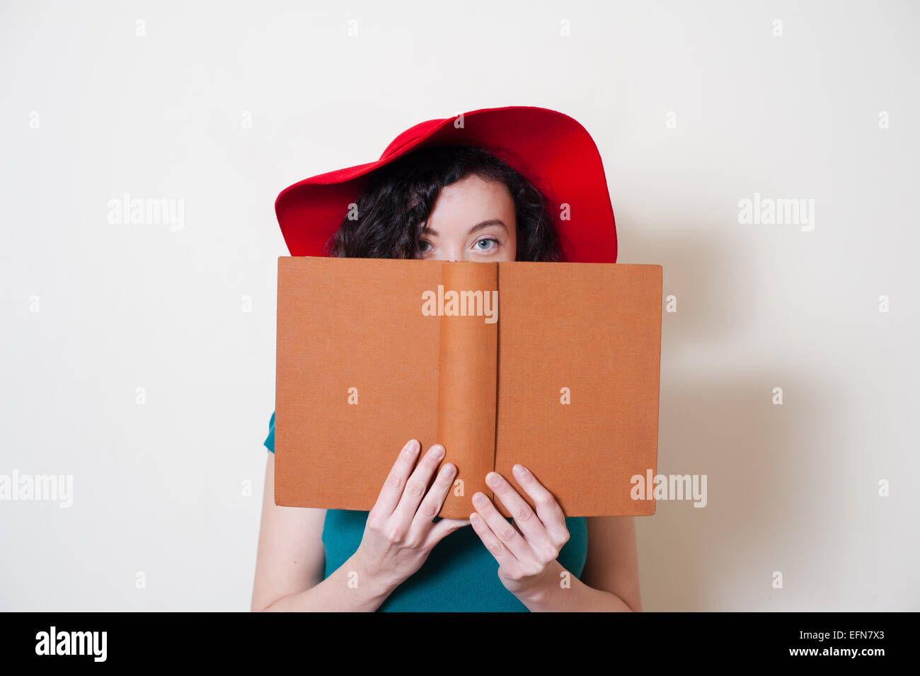 Jeune femme avec red hat holding book partiellement couvert visage semble derrière sur fond blanc Banque D'Images
