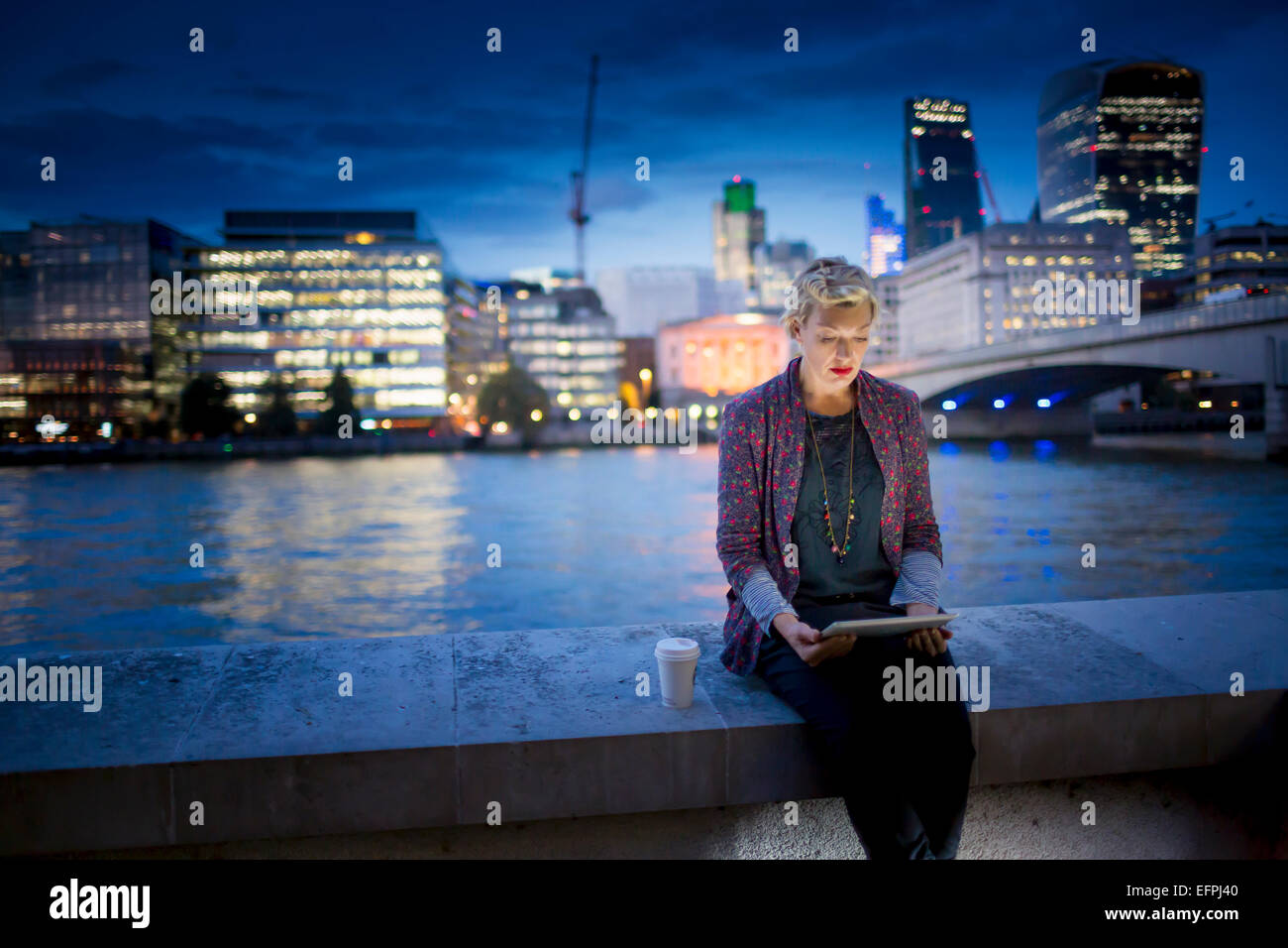 Young businesswoman sitting on Thames waterfront using digital tablet at night, London, UK Banque D'Images