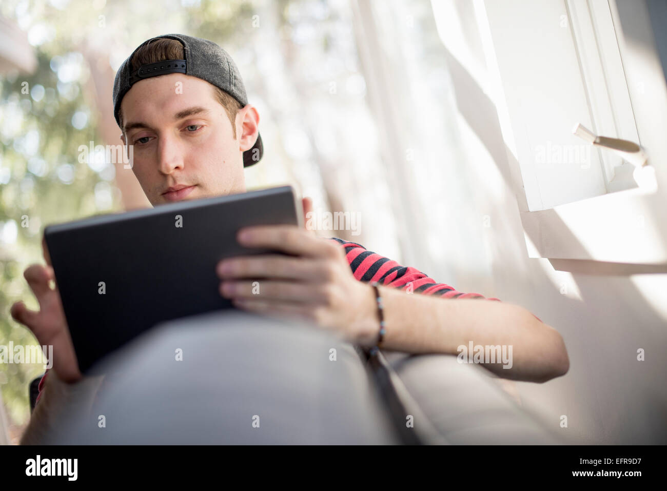Homme portant une casquette de baseball en arrière, assise sur un canapé, regardant une tablette numérique. Banque D'Images