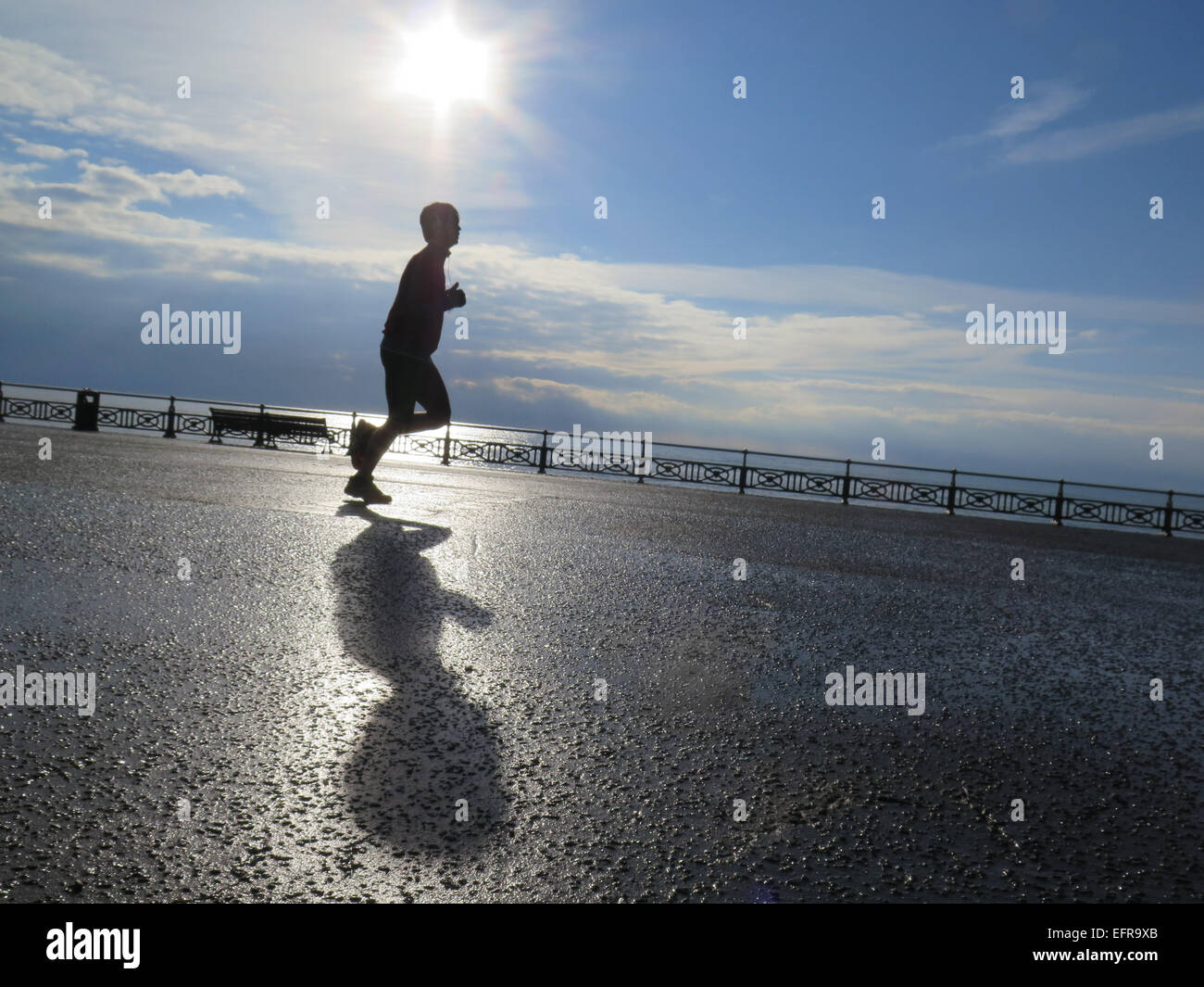 Un homme jogger court le long de la promenade de pelouses Hove casting son ombre dans la lumière du soleil tôt le matin au bord de la mer scintillante. Banque D'Images