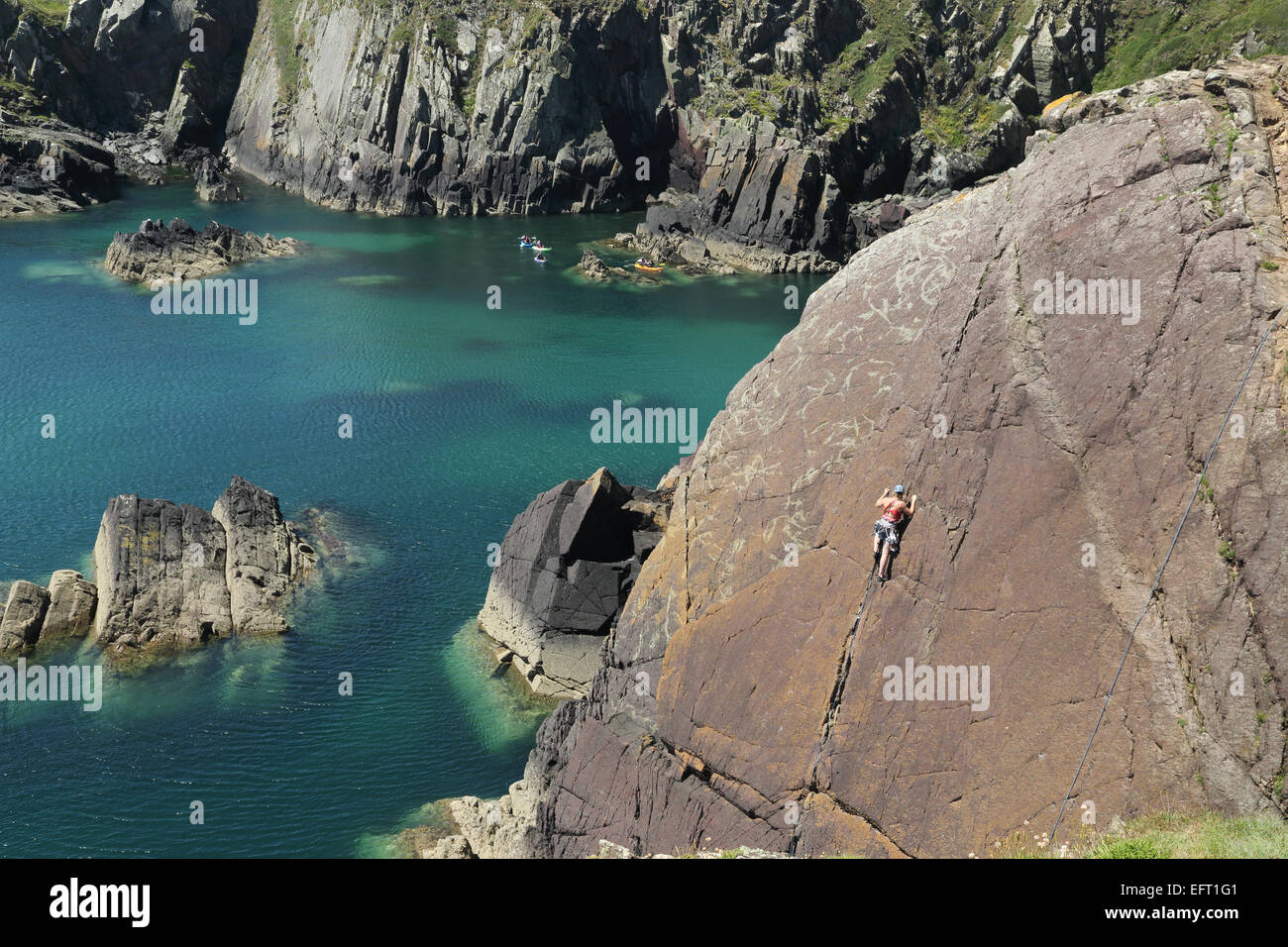 Female rock climber on coastal rock face off Pembrokeshire Coastal Path avec les canoéistes en arrière-plan Banque D'Images