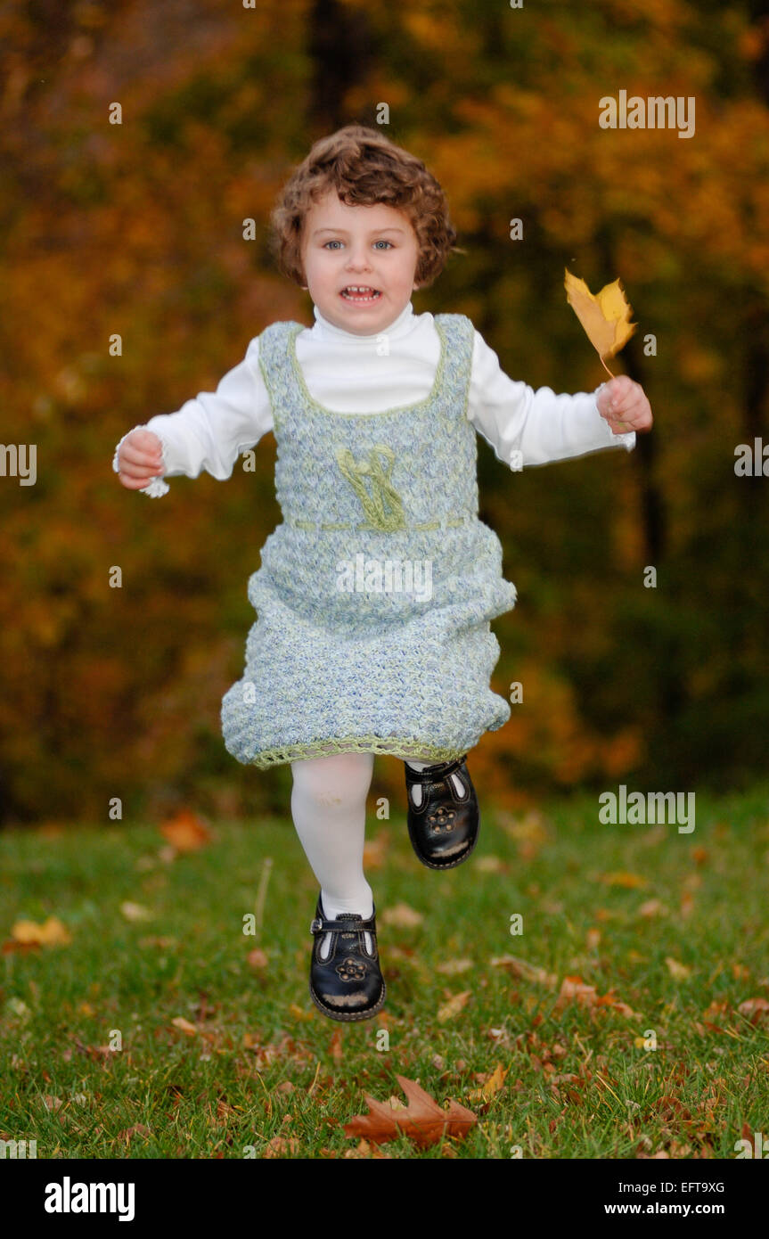 Jeune fille jouant avec les feuilles d'automne sur la pelouse verte à l'extérieur portant une jolie robe au crochet Banque D'Images