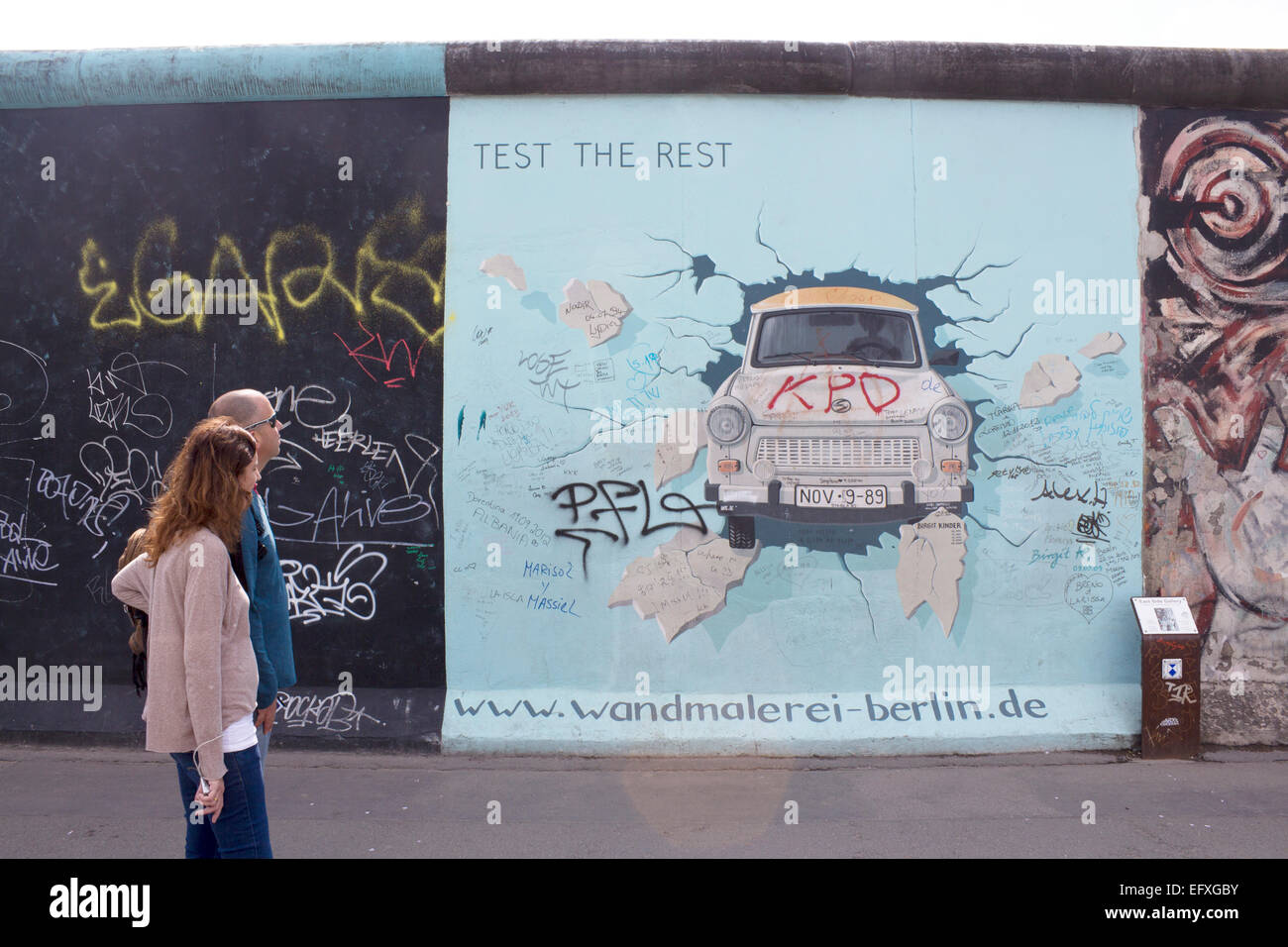 Couple walking passé Birgit Kinder's artwork de voiture Trabant éclatant à travers la paroi Cinquième mise à jour de l'artwork avec graffit supplémentaires Banque D'Images