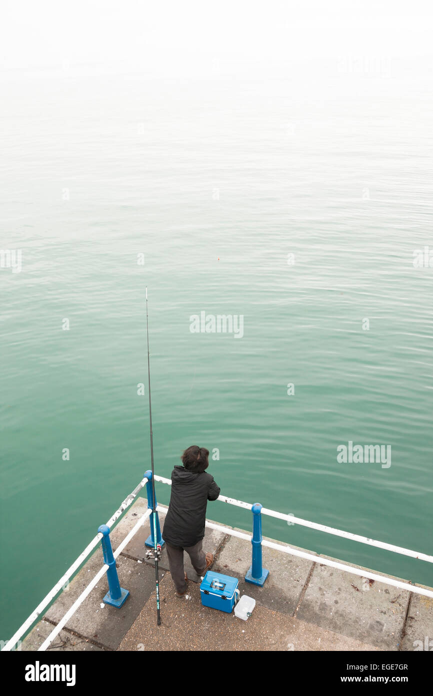 Lone patient pêcheur en manteau noir à l'extrémité de tige reposant sur la jetée rambarde en attente d'une morsure avec boîte bleue Banque D'Images