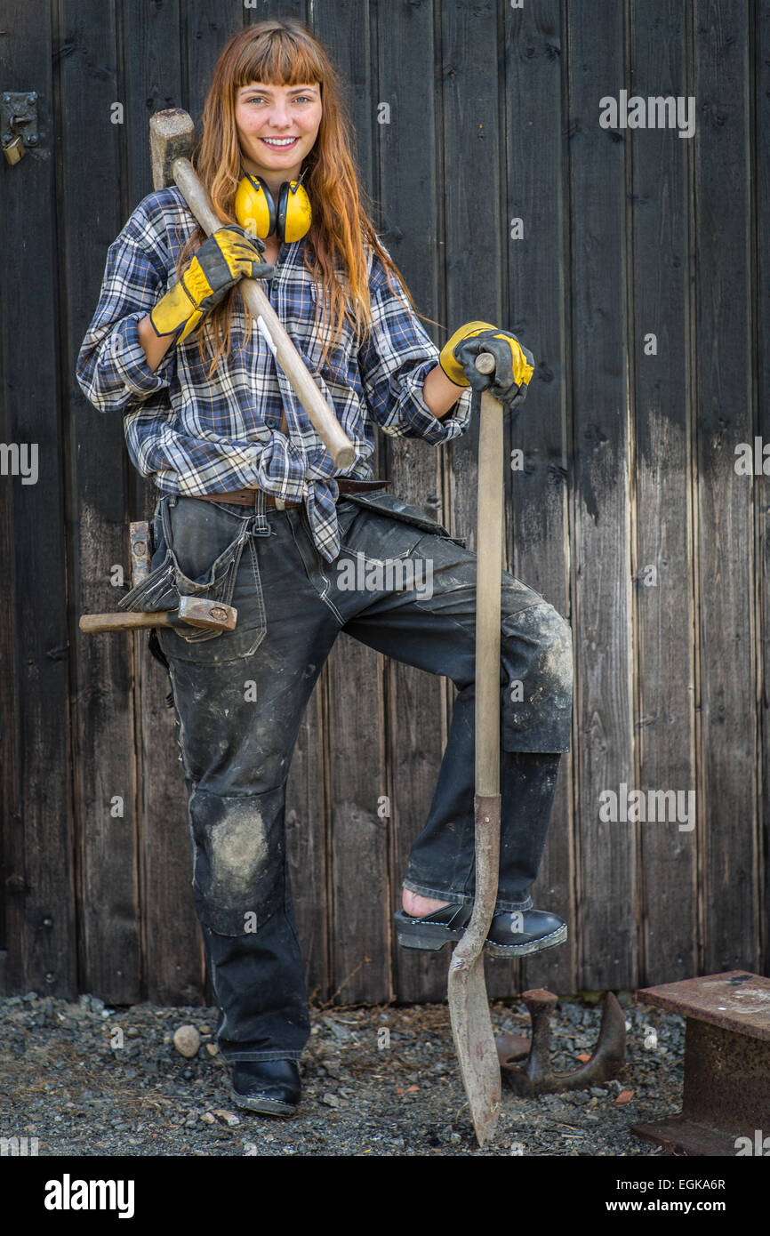 Girl in construction worker clothing, debout avec une pelle et un marteau en face d'une grange Banque D'Images