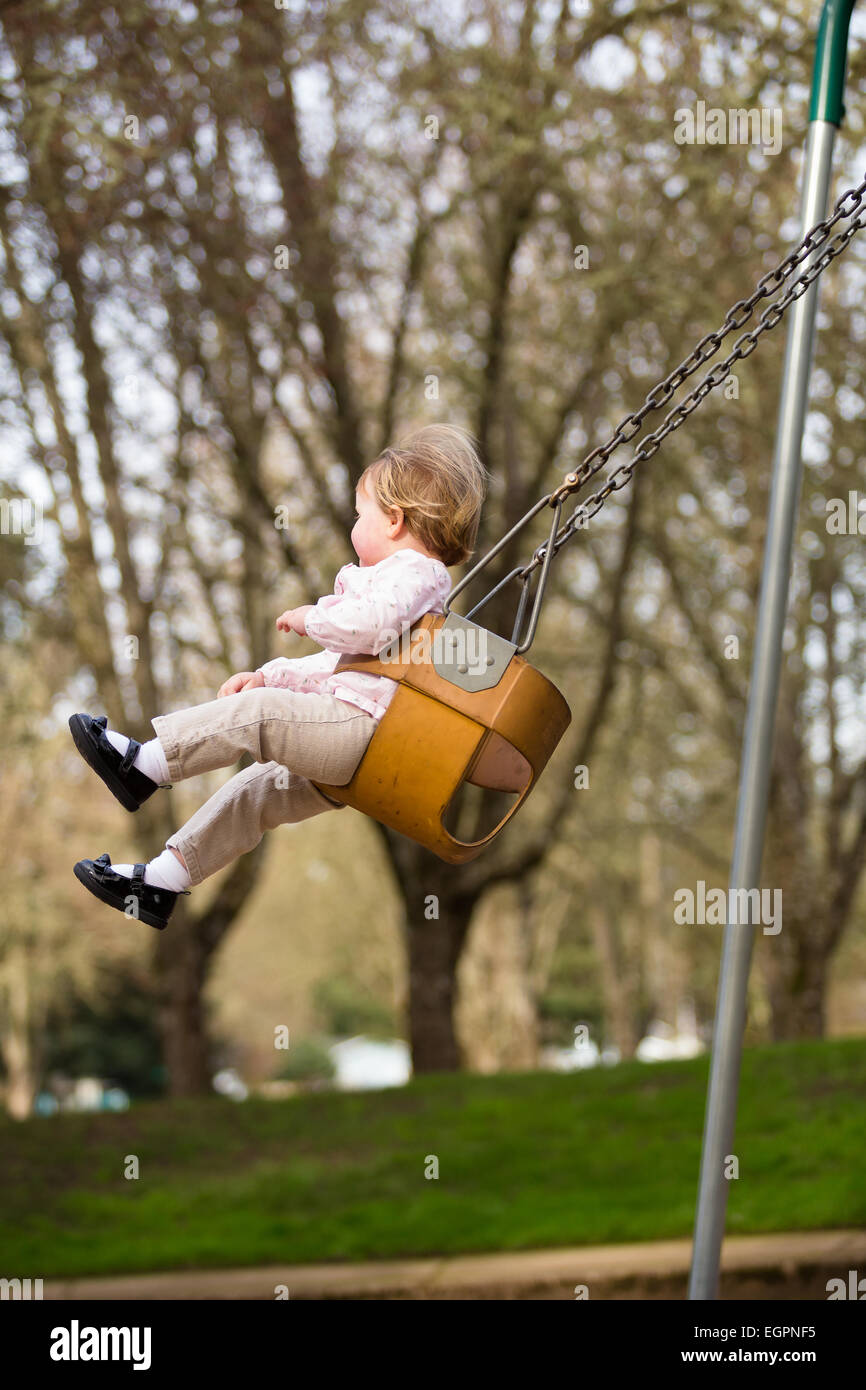 Un an fille sur une balançoire dans un parc dans la lumière naturelle. Banque D'Images